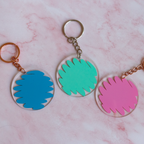 Three colorful keychains with scribble designs on a pink marble background